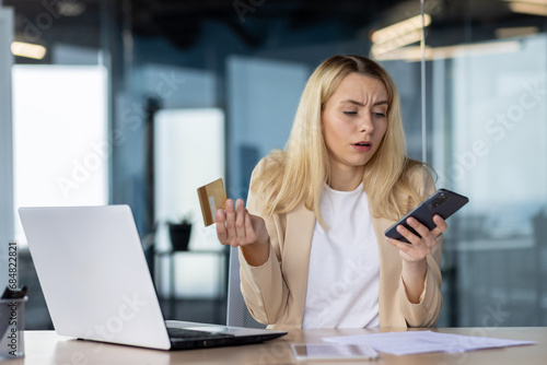 Frustrated sad upset woman working inside office, cheated business woman refused online money transfer, female worker displeased holding bank credit card and phone
