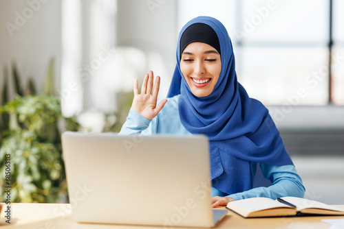 Joyful young muslim woman entrepreneur smiling and gesticulating while sitting at desk and talking to online coworkers during online meeting