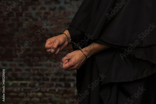 The hands of a monk or priest in handcuffs on the background of a stone wall. Concept: old criminal, criminal liability, religious figure.