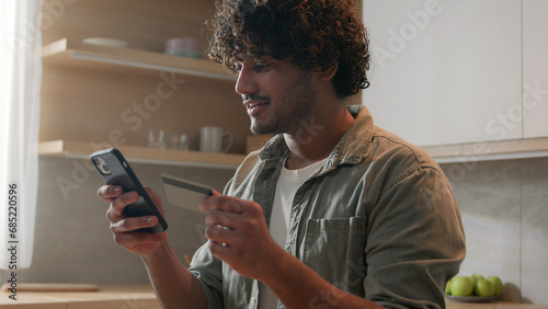 Happy Arabian man with phone and credit card using instant mobile payment at home kitchen Indian guy male customer shopper buyer buying online purchase smartphone shopping food delivery e-banking app