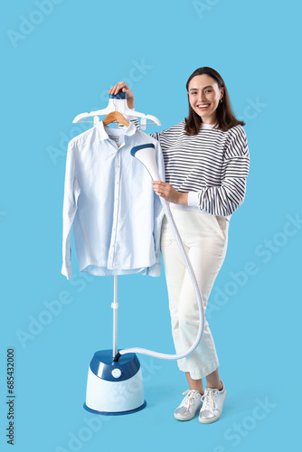 Pretty young woman with modern steamer and clothes on blue background