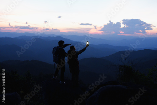 Silhouette of Asian two male standing raised hands with trekking poles and kerosene black lamp on cliff edge on top of rock mountain with at sunset rays over the clouds background,