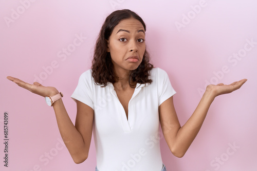 Young hispanic woman wearing casual white t shirt clueless and confused expression with arms and hands raised. doubt concept.