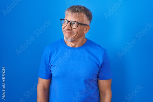 Hispanic man with grey hair standing over blue background smiling looking to the side and staring away thinking.