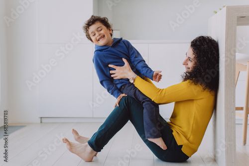 Mom playing with son at home. Cheerful little boy happy to spend time with mother on weekend. Family funny moment. Positive babysitter entertains toddler, tickling him, smiling, having fun.