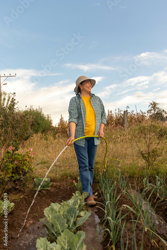 Mujer latina adulta caminando con una manguera llegando las hortalizas en medio de su huerta foto contrapicada que demuestra el empoderamiento femenino 