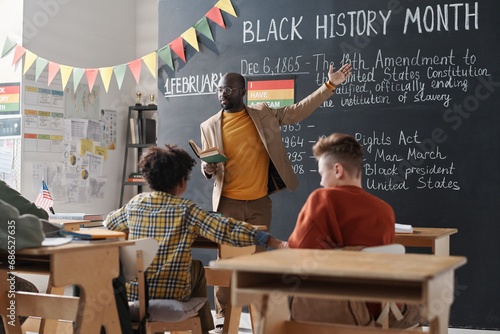 African AMerican teacher pointing at blackboard and talking about Black History Month to school children during a lesson