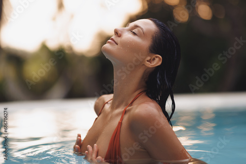 Tropical paradise, luxury relaxation of bikini brunette woman in sexy orange swimsuit in infinity pool at sunset covering her breast by hands. High quality portrait photo