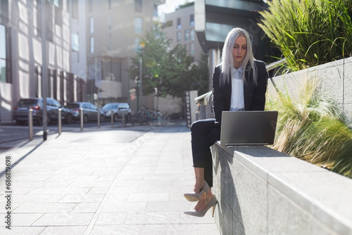 Young businesswoman sitting on a wall in the city using laptop