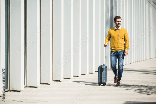 Young man with rolling suitcase and earphones in the city on the go