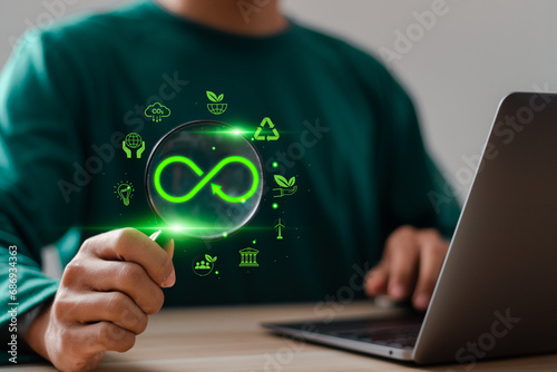 Businessman use laptop and hand holding magnifying glass focus to Infinity symbol for circular economy to reduce waste by reusing, Reduce pollution for future business and environmental growth.