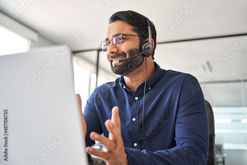 Happy Indian call center agent wearing headset talking to client working in customer support office. Professional contract service telemarketing operator using laptop having conversation. Candid shot