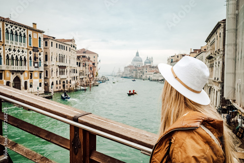 A stylish woman gazes upon the bustling cityscape and tranquil river, adorned in a fashionable hat and clothing, as a ship and gondola glide by on the water