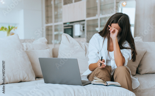 tired brunette is sitting on the couch at home working with a laptop and with her diary open, she covers her face and touches her forehead, exhausted, Crying lady, grief.