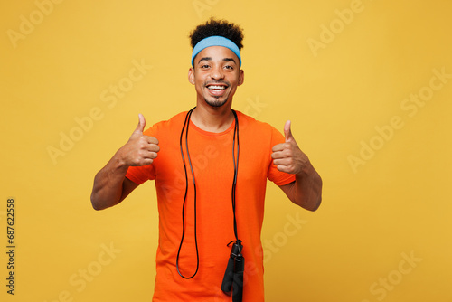 Young fitness trainer instructor sporty man sportsman wears orange t-shirt hold skipping rope show thumb up spend time in home gym isolated on plain yellow background. Workout sport fit abs concept.