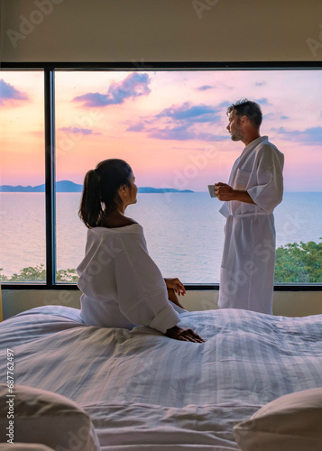 couple man and woman in front of the bed in a hotel bedroom during sunrise drinking a cup of coffee, a European man and an Asian woman in a hotel room during sunset in Thailand