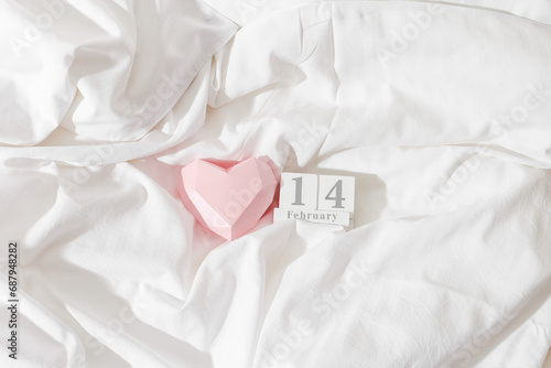 Valentine's Day concept, pink paper valentine heart and 14 february holiday date on wooden calendar in bed on on white crumpled sheets. Minimal style flat lay photo, top view, copyspace, pastel