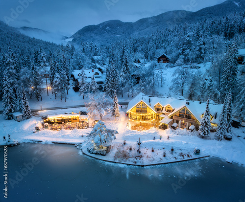 Aerial view of illuminated houses in fairy village in snow, forest, Jasna lake, street lights at winter night. Top view of alpine mountains in fog, snowy pine trees at dusk. Kranjska Gora, Slovenia