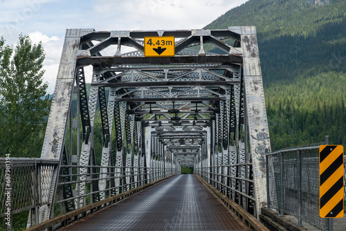 Drivers perspective of a steel truss bridge across the river in mountainous area of Canada with bridge deck of steel grating and signs indicating maximum height