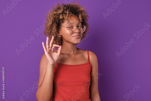 Young beautiful confident African American woman student with glasses shows confirming gesture saying that everything is ok or agreeing to your commercial proposal stands posing on purple background.