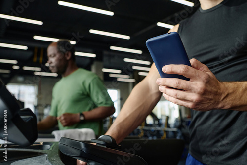 Unknown caucasian guy training at gym holding smartphone, elderly black man exercising on background