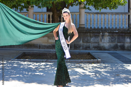 Pretty young woman winner of a beauty pageant dressed in a green sequined dress. Young woman wears diamond crown and winner's sash and poses for photo. Fashion and beauty concept.