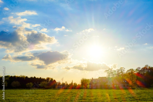 This image captures the serene beauty of a countryside sunset, with the sun's golden rays piercing through the clouds and casting a warm glow over the expansive field. The rays create a dramatic flare