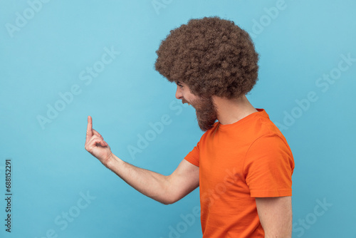 Side view of angry man with Afro hairstyle in orange T-shirt showing middle finger and asking to get off expressing negativity, disrespectful behaviour. Indoor studio shot isolated on blue background.