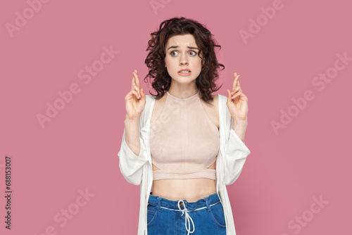Portrait of hopeful attractive woman with curly hair wearing casual style outfit crossing her finger for good luck, waiting for miracle. Indoor studio shot isolated on pink background.
