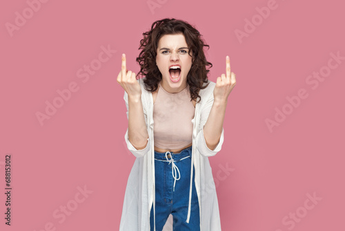 Portrait of rude impolite woman with curly hair wearing casual style outfit standing showing middle fingers, screaming with hate and anger. Indoor studio shot isolated on pink background.