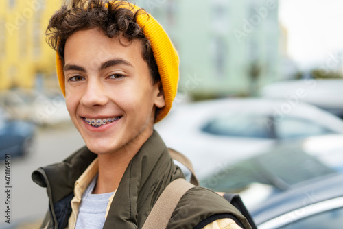 Portrait attractive smiling millennial boy wearing yellow hat, with dental braces on teeth looking at camera on urban street