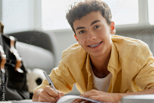 Portrait of attractive smiling boy with dental braces studying, learning language, taking notes lying on floor looking at camera. Education concept