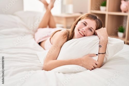 Young blonde woman hugging pillow lying on bed smiling at bedroom