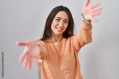 Young brunette woman standing over white background looking at the camera smiling with open arms for hug. cheerful expression embracing happiness.