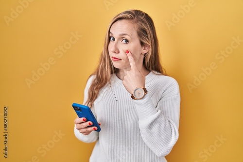 Young blonde woman using smartphone typing message pointing to the eye watching you gesture, suspicious expression