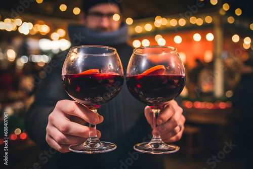 Two glasses of hot red mulled clink glasses with orange in hands. Christmas market in the city park. Rustic decor, cozy atmosphere, festive mood.