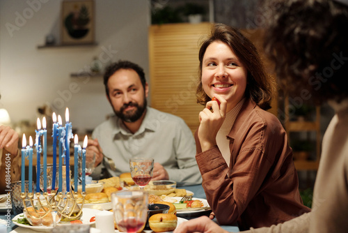 Young smiling woman looking at her husband by served festive table with homemade food and drinks while listening to him during discussion