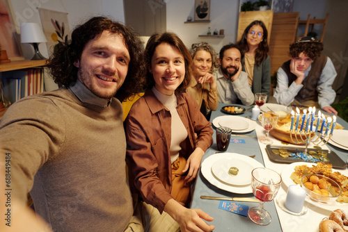 Happy young couple taking selfie with members of their family while sitting by served festive table and enjoying Hanukkah dinner