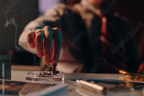 Close-up of hand of young man stubbing butt of cigarette in ashtray standing on table among business and office supplies