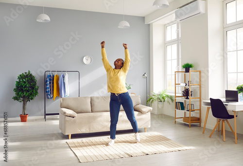 Happy cheerful young African American woman in casual clothes enjoying free time at home, standing in living room with sofa, raising hands up, stretching, smiling and charging with positive energy