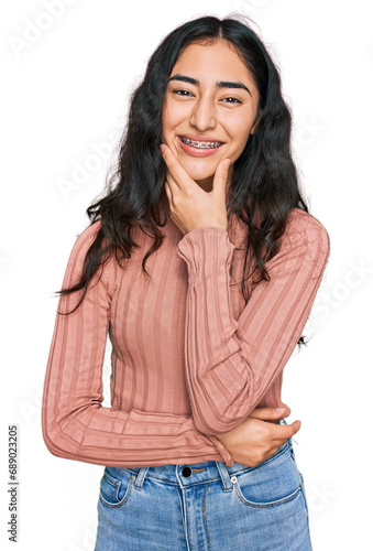 Hispanic teenager girl with dental braces wearing casual clothes looking confident at the camera smiling with crossed arms and hand raised on chin. thinking positive.