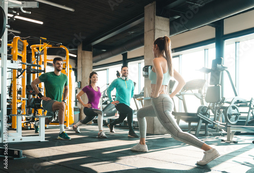 Group of athletic people exercising in a health club. Cheerful young athletes doing lunges in gym under supervision of their coach. Female fitness instructor stretching with a group of three people.