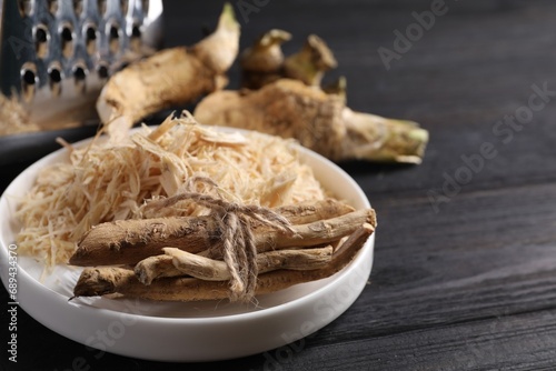 Plate with whole and grated horseradish roots on black wooden table, closeup. Space for text