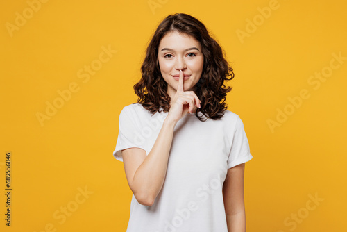 Young secret smiling Caucasian woman she wearing white blank t-shirt casual clothes saying hush be quiet with finger on lips shhh gesture isolated on plain yellow orange background. Lifestyle concept.