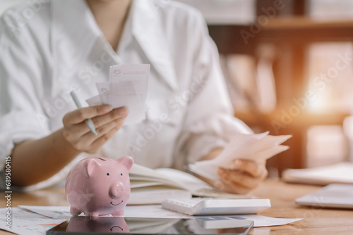 Woman sitting at desk managing expenses, calculating expenses, paying bills using laptop online, making household financial analysis, closer focus on the pink piggy bank.