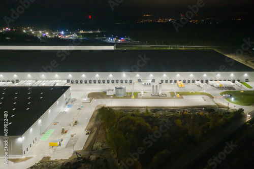 Centrum logistyczne w nocnych ciemnościach. Teren wokół jak i budynki oświetlone są sztucznym światłem elektrycznym. Zdjęcie zrobione przy użyciu drona.