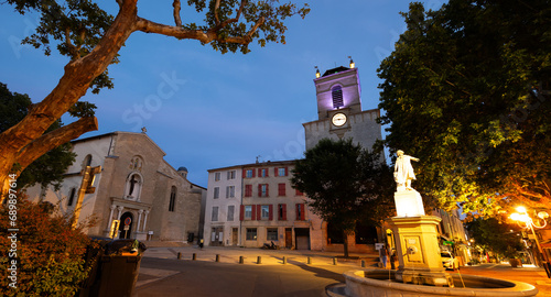 Historic square Place Mirabeau in French town of Pertuis overlooking illuminated statue with fountain on background with medieval donjon with clock and parish church of Saint-Nicolas on summer evening