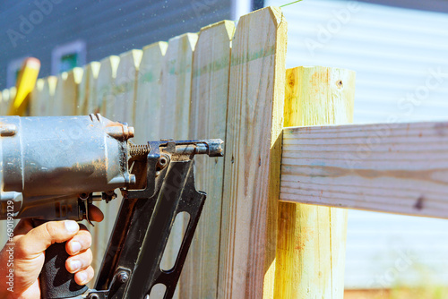 By using an air nail gun, he constructs sections of wooden fence around his yard