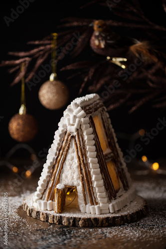 Handmade gingerbread A-form house for Christmas and New Year with icing sugar snow on a dark background with decorations and Christmas lights.