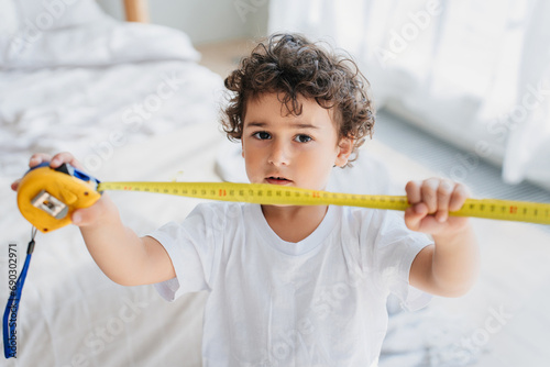 Curly confused handsome little boy holding measuring roulette, wants growing faster. Children psychology. Pretty kid puzzled by his small height. Spanish kid with tape measuring at home showing height
